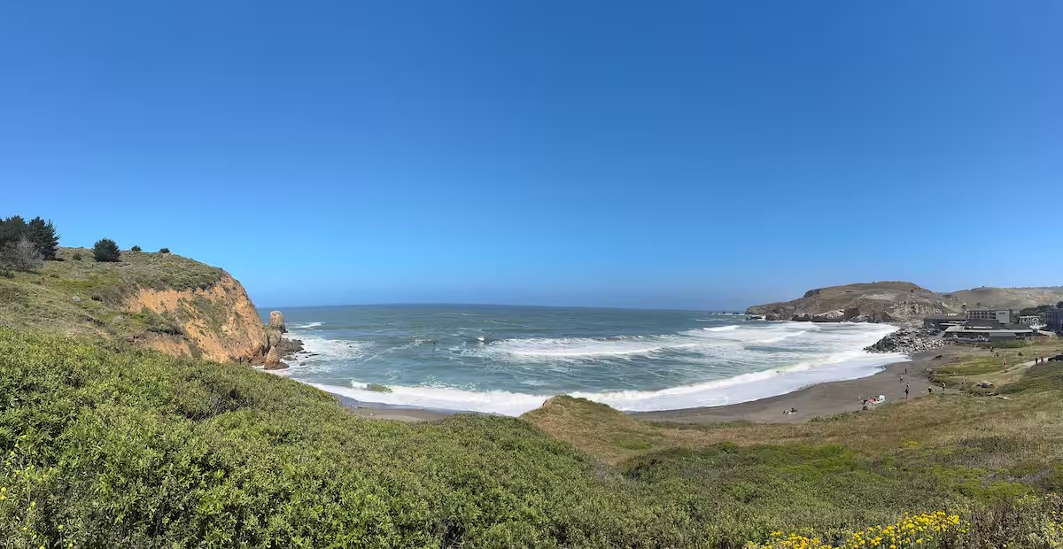 Wide view of a Pacifica cove with cliffs, green scrub, and whitecaps breaking on the beach